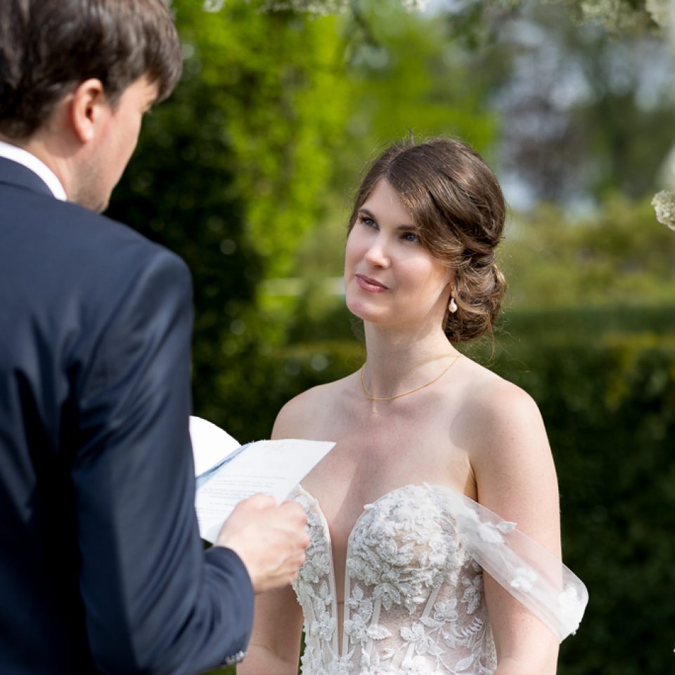 Bride in a flowing white dress stood attentively in front of the groom in a lush castle garden, listening to him read vows during their wedding ceremony.